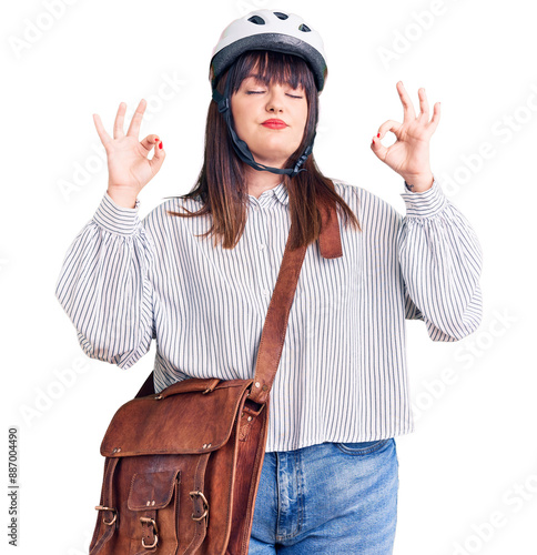 Young plus size woman wearing bike helmet and leather bag relax and smiling with eyes closed doing meditation gesture with fingers. yoga concept. photo