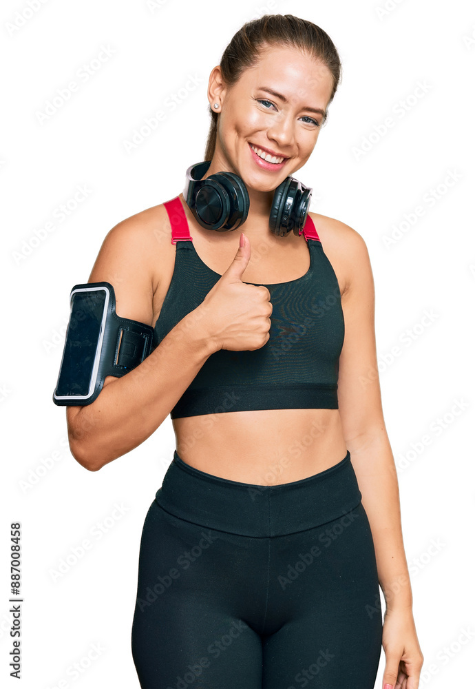Beautiful blonde woman wearing gym clothes and using headphones doing happy thumbs up gesture with hand. approving expression looking at the camera showing success. Beautiful blonde woman wearing gym clothes and using headphones doing happy thumbs up gesture with hand. approving expression looking at the camera showing success.