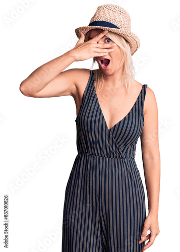 Young beautiful blonde woman wearing summer hat and dress peeking in shock covering face and eyes with hand, looking through fingers with embarrassed expression.