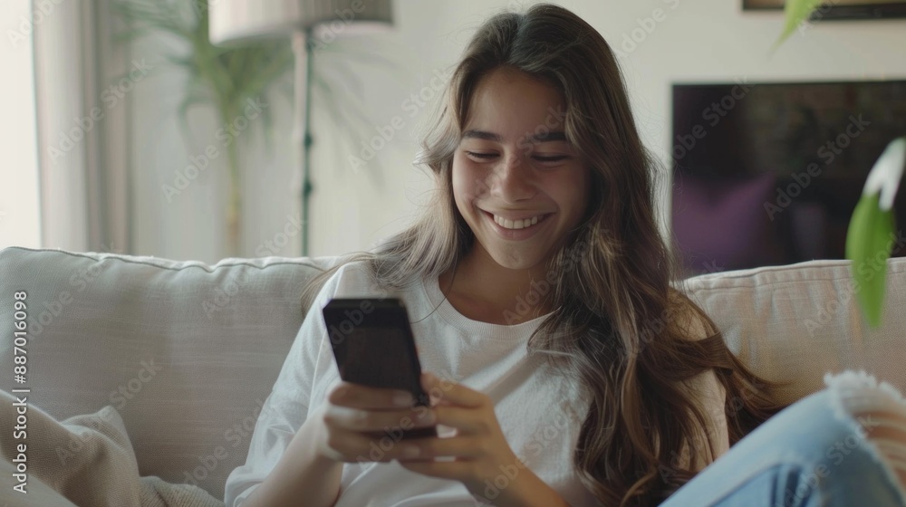 A young woman with long brown hair, wearing a white t-shirt and blue jeans, smiles as she sits on a white couch and looks at her phone A young woman with long brown hair, wearing a white t-shirt and blue jeans, smiles as she sits on a white couch and looks at her phone