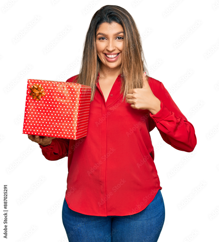 Beautiful brunette woman holding gift smiling happy and positive, thumb up doing excellent and approval sign Beautiful brunette woman holding gift smiling happy and positive, thumb up doing excellent and approval sign