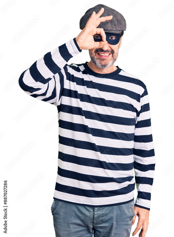 Middle age handsome man wearing burglar mask doing ok gesture with hand smiling, eye looking through fingers with happy face. Middle age handsome man wearing burglar mask doing ok gesture with hand smiling, eye looking through fingers with happy face.