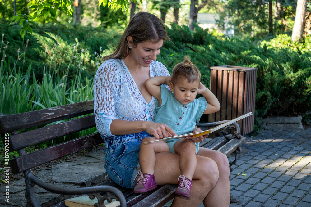 The mother is sitting with her baby daughter on a bench in the park and reading a book together. Child development and education. The mother is sitting with her baby daughter on a bench in the park and reading a book together. Child development and education.