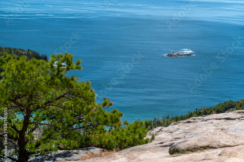 Acadia National Park View From The Summit. A panoramic view of the ocean from the summit of a rocky mountain, with a small island in the distance. Lush green trees and shrubs surround the rocky peak,