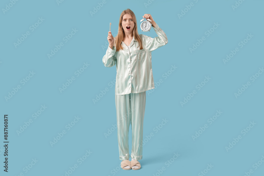 Shocked young woman in pajamas with alarm clock and toothbrush on blue background Shocked young woman in pajamas with alarm clock and toothbrush on blue background