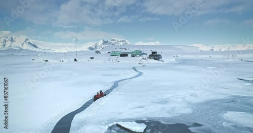 Scientist from polar station base riding zodiac boat in melted steam. Frozen ocean ice, mountains and science buildings in background. South Pole tourism, travel, expedition. Aerial drone panorama