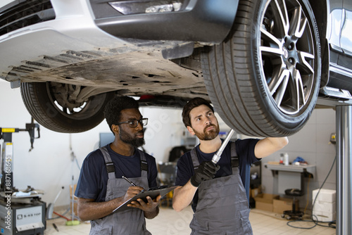 Focused automotive technicians working on car maintenance and safety inspection. Two mechanics check vehicle chassis on lift in professional auto repair shop. photo