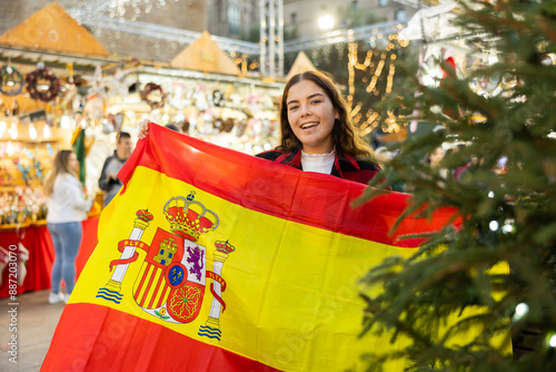 Cheerful young girl holding national flag of Spain, standing outdoors against blurred background of illuminated shopping stalls at traditional Christmas city market..