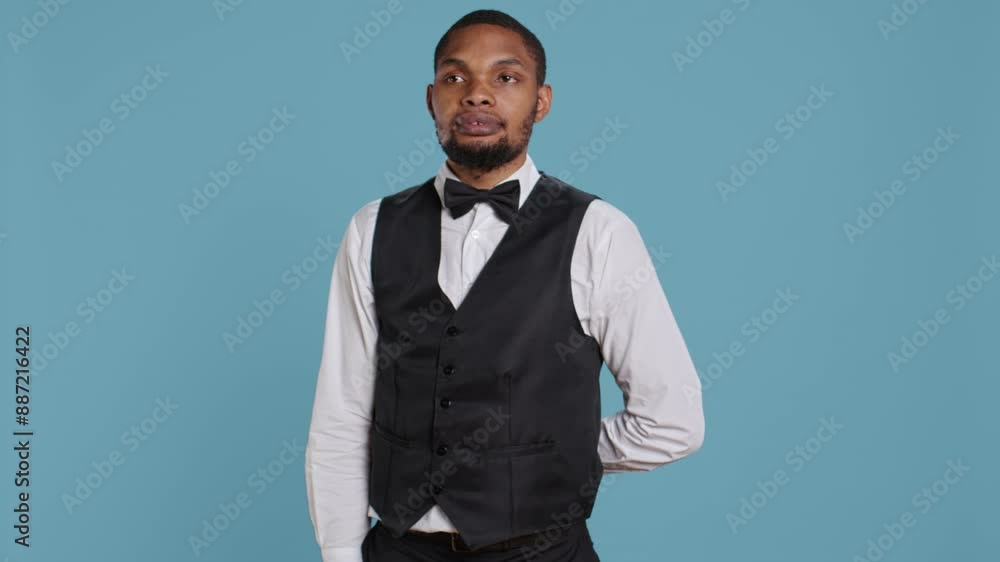 Bellboy hotel porter welcoming guests at the five star lodging resort, smiling and greeting people against blue background. Employee wearing a tuxedo and tie, tourism industry. Camera B. Bellboy hotel porter welcoming guests at the five star lodging resort, smiling and greeting people against blue background. Employee wearing a tuxedo and tie, tourism industry. Camera B.