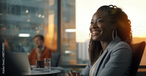 Charming black woman in office, dressed in suit. She sits at a stylish modern desk and smiles sweetly. Diversity concept.