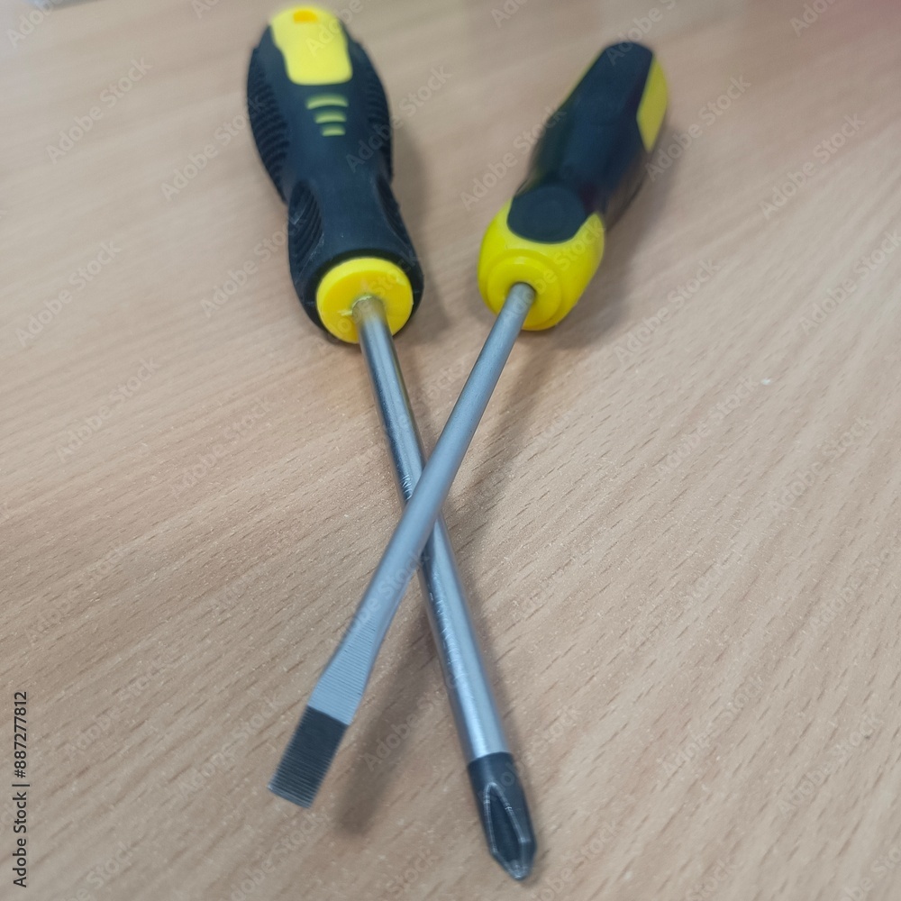 Two screwdrivers on a wooden surface, one with a flathead tip and the other with a Phillips head. Both have black handles with yellow accents, and their metal shafts cross each other. Two screwdrivers on a wooden surface, one with a flathead tip and the other with a Phillips head. Both have black handles with yellow accents, and their metal shafts cross each other.