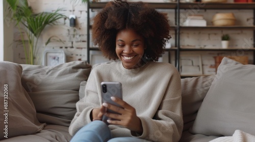 A young woman with curly hair sits comfortably on a sofa, her eyes fixed on her phone. She smiles radiantly as she reads a text or watches a video