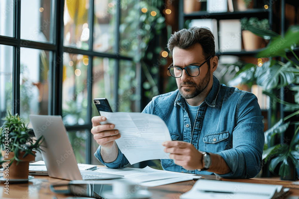 Focused Casual Entrepreneur Accountant in Glasses Analyzing Documents at Desk with Laptop Near Window Focused Casual Entrepreneur Accountant in Glasses Analyzing Documents at Desk with Laptop Near Window