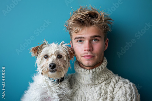 Young man with messy blonde hair is posing with a small white dog on a blue background, owner and dog look alike. 