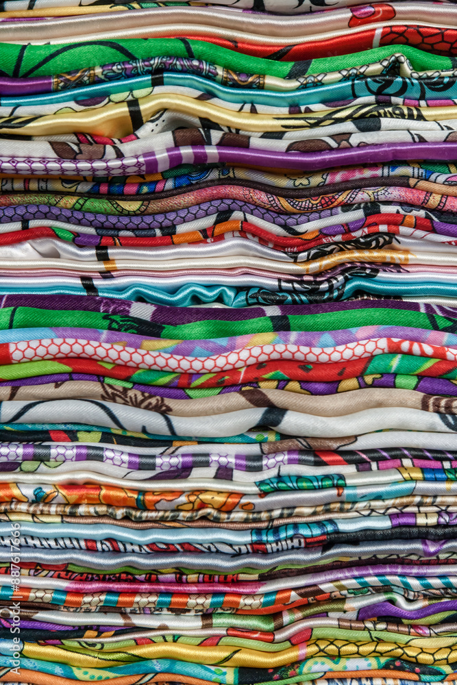 Fototapeta premium Stack of colorful, patterned fabrics, traditional shawls, arranged in layers in a local market, featuring various designs and vibrant colors, creating a visually striking and textured appearance.