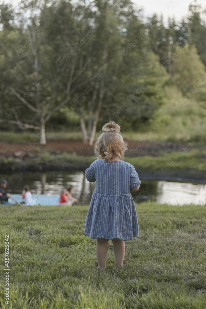 Toddler Standing in Grass Overlooking a Pond with a Boat Toddler Standing in Grass Overlooking a Pond with a Boat