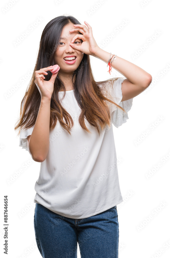 Young asian woman eating pink macaron sweet over isolated background with happy face smiling doing ok sign with hand on eye looking through fingers Young asian woman eating pink macaron sweet over isolated background with happy face smiling doing ok sign with hand on eye looking through fingers