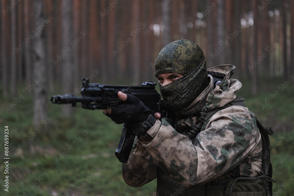 A angry soldier in balaclava and camouflage takes aim with an AK 12 rifle in the forest at dusk. A angry soldier in balaclava and camouflage takes aim with an AK 12 rifle in the forest at dusk.