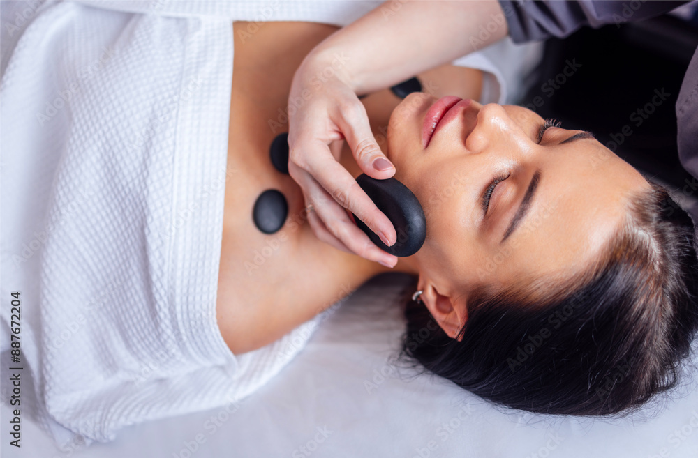 Close-up of a portrait of a young woman with her eyes closed receiving a facial massage with hot stones. Close-up of a portrait of a young woman with her eyes closed receiving a facial massage with hot stones.