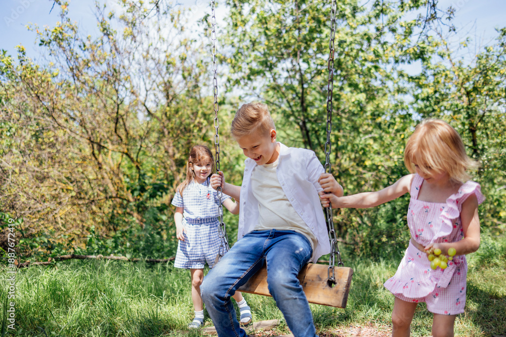 Fototapeta premium Pretty little sisters in summer is rocking brother on swing.