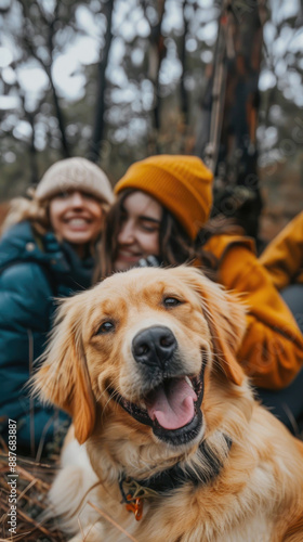 A golden retriever dog smiles happily at the camera while two friends sit behind him in the woods © sommersby