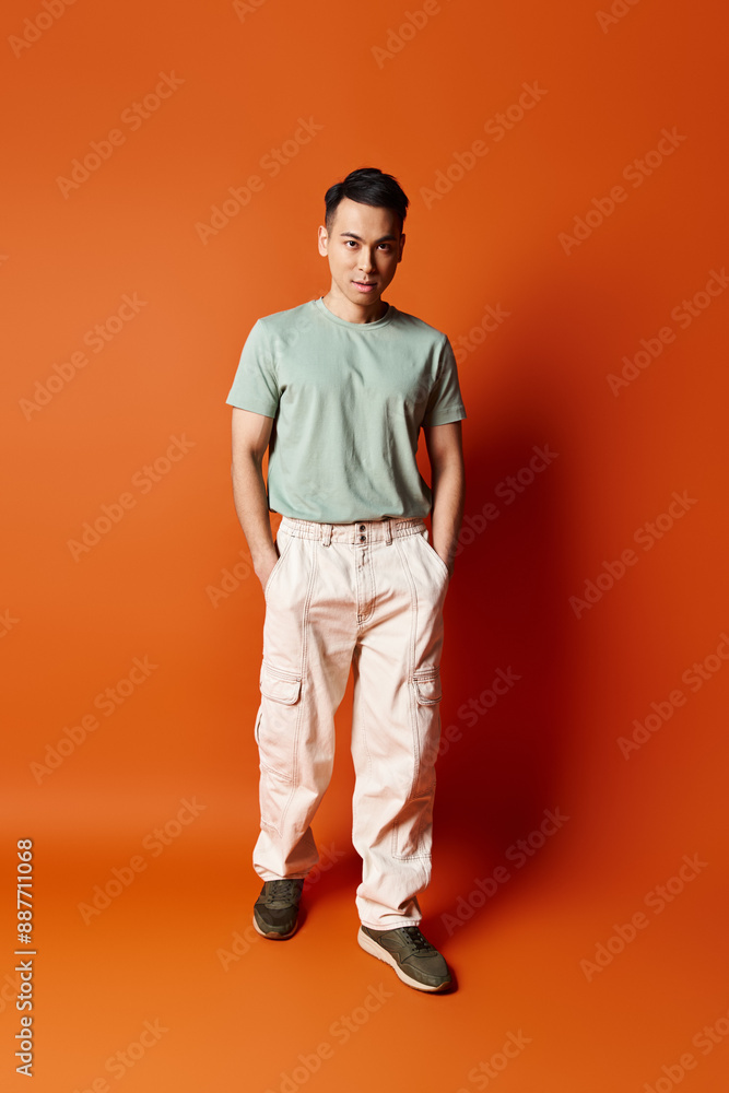 Fototapeta premium A handsome Asian man stands confidently in front of a vibrant orange wall, emanating a sense of style and individuality.