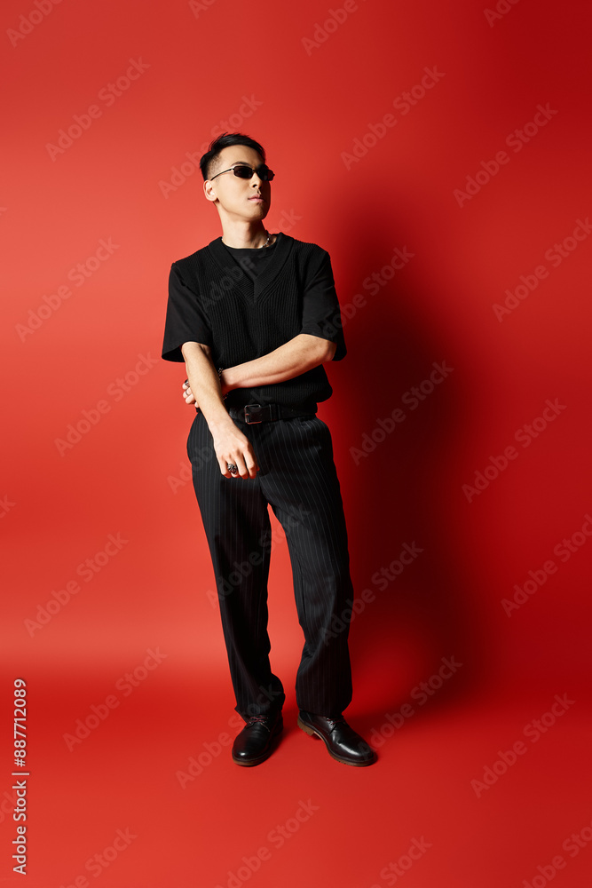 A stylish Asian man, dressed in black attire, stands confidently in front of a bold red background in a studio setting. A stylish Asian man, dressed in black attire, stands confidently in front of a bold red background in a studio setting.