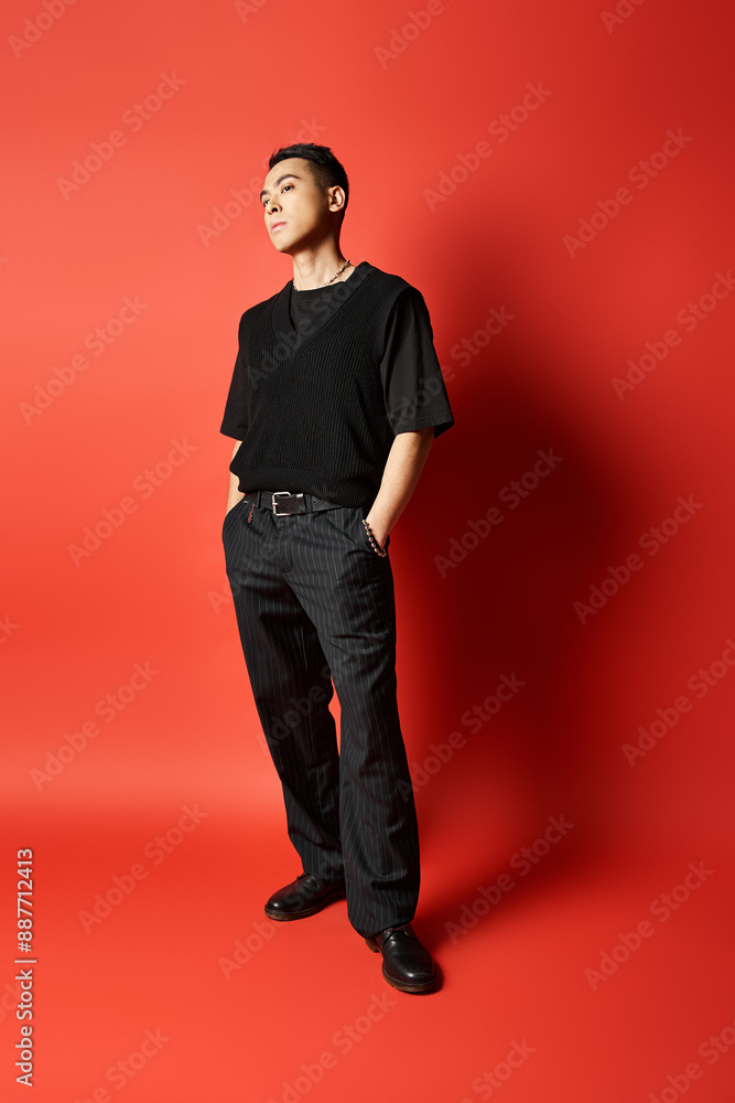 A fashionable Asian man in black attire stands confidently in front of a striking red wall in a studio setting. A fashionable Asian man in black attire stands confidently in front of a striking red wall in a studio setting.