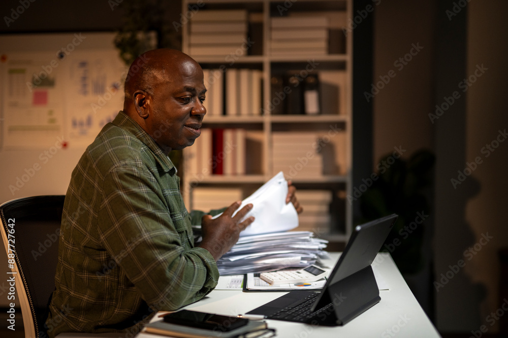 Fototapeta premium A man is sitting at a desk with a laptop and a stack of papers
