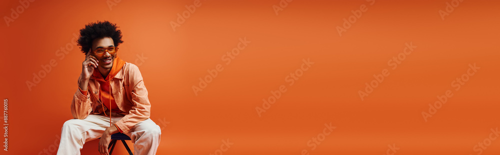 Fototapeta premium Curly African American man, stylishly dressed, sitting on chair on orange background.