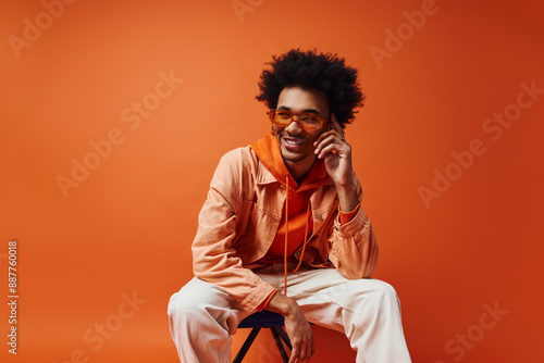 Konstfotografi A trendy African American man with curly hair, sunglasses, and stylish attire sits on a chair against an orange background