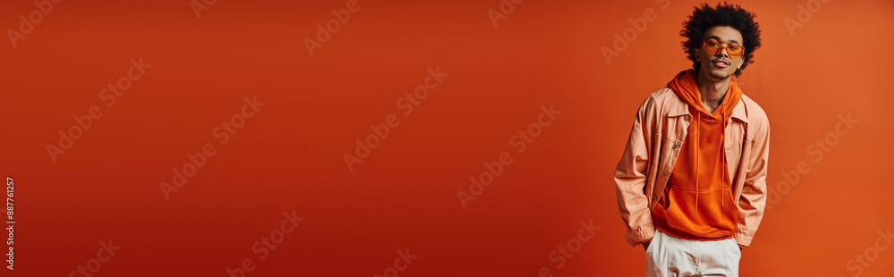 A stylish young African American man, wearing sunglasses and a trendy outfit, poses in front of a bright red wall.