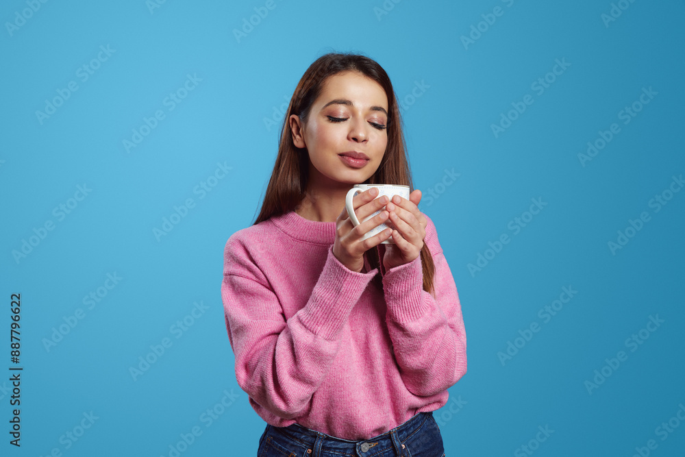 Portrait of satisfied hispanic girl holding cup of hot coffee with closes eyes and enjoying aroma, wearing pink jumper, isolated on blue background