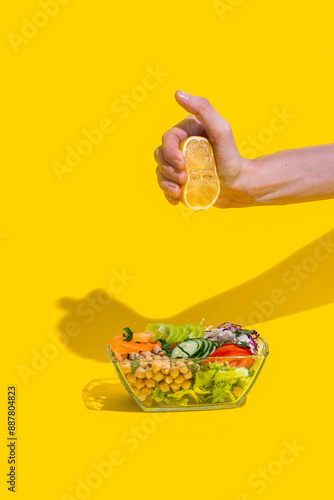 Woman squeezing fresh lemon over bowl with salad on a jellow background