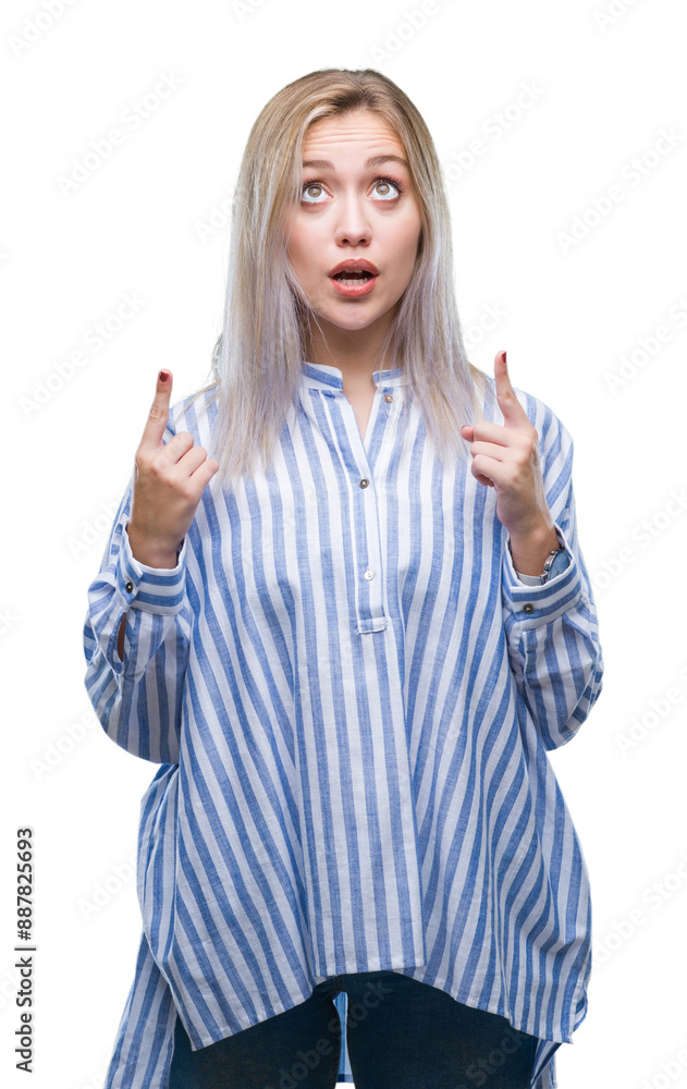Young blonde woman over isolated background amazed and surprised looking up and pointing with fingers and raised arms. Young blonde woman over isolated background amazed and surprised looking up and pointing with fingers and raised arms.