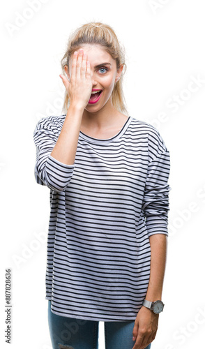 Young beautiful blonde woman wearing stripes sweater over isolated background covering one eye with hand with confident smile on face and surprise emotion.