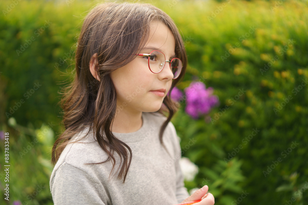 A lonely girl with glasses with curly hair in the backyard in the summer writes intently into a notebook with a pencil. Concept of children having fun outdoors. A lonely girl with glasses with curly hair in the backyard in the summer writes intently into a notebook with a pencil. Concept of children having fun outdoors.