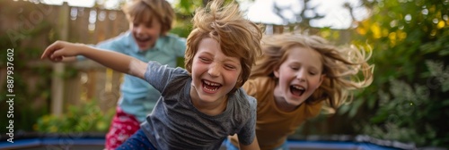 Three children, two boys and one girl, jump and laugh on a trampoline in a backyard. The two children in the foreground are looking directly at the camera, smiling © Elmira