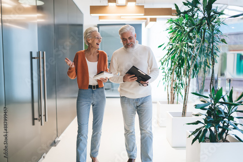 elderly Caucasian man and woman, both with white hair, walk through modern office hallway. Mature business colleagues engaged in cheerful discussion,  professional and friendly work environment © BullRun