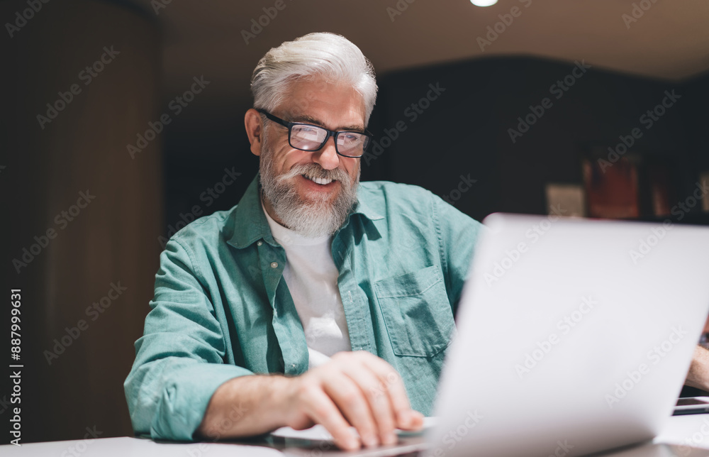 Fototapeta premium Senior man wearing glasses, working a laptop in coworking. Businessman focused and content while analyzing documents. Nighttime office environment, emphasizing concentration and financial dedication