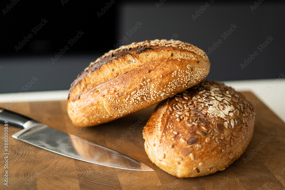 Two loaves of sourdough bread, freshly baked and still warm, are stacked on a wooden surface. The loaves are covered in seeds sesame and sunflower, adding a rustic and flavorful touch. Two loaves of sourdough bread, freshly baked and still warm, are stacked on a wooden surface. The loaves are covered in seeds sesame and sunflower, adding a rustic and flavorful touch.