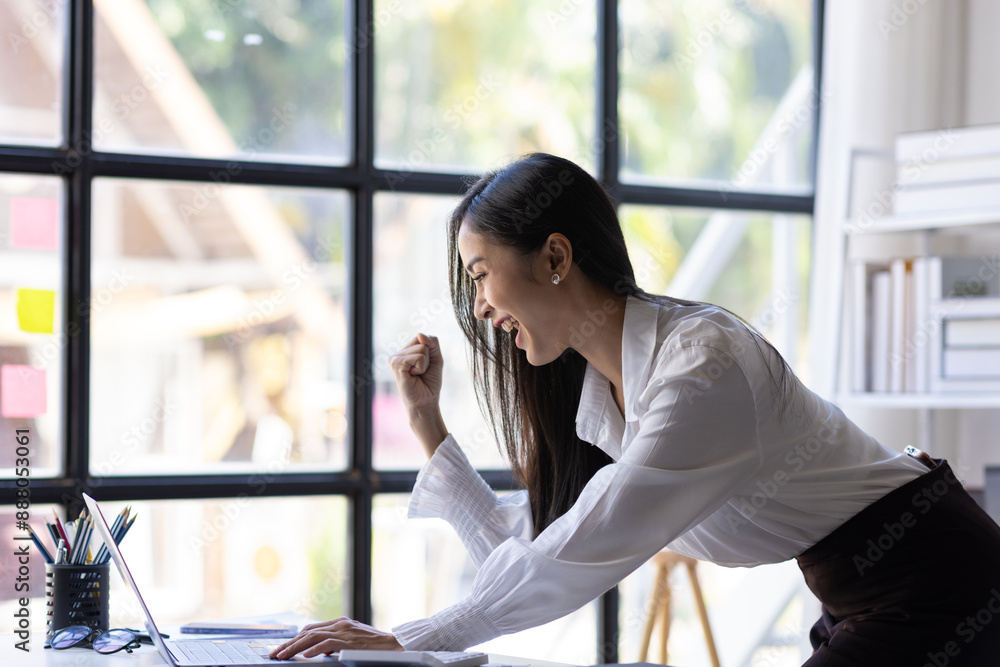Fototapeta premium Successful Asian businesswoman happy looking at laptop and excited. Woman in office looking at laptop surprised to receive bonus and promotion.