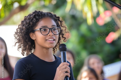 A confident young woman with glasses is speaking into a microphone at an outdoor event, engaging with the audience. women's speaking, Womens Equality Day