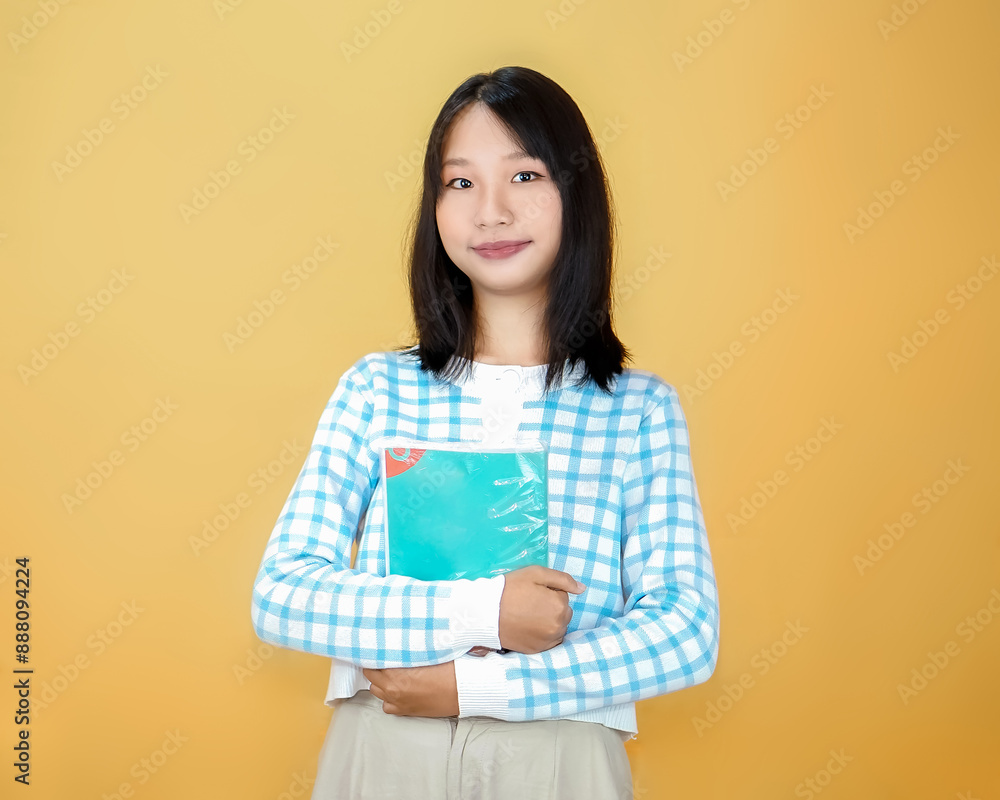 Fototapeta premium young asian woman looking at the camera with a book in her hand isolated on a yellow background
