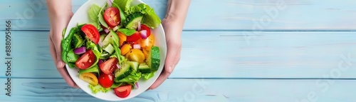 Hands holding a fresh, colorful salad bowl with tomatoes, greens, and cucumbers over a light blue wooden table surface.