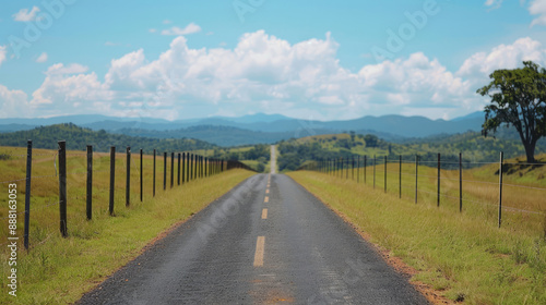 Straight country road stretching into the horizon under a blue sky with scattered clouds, surrounded by green pastures and a distant mountain range.
