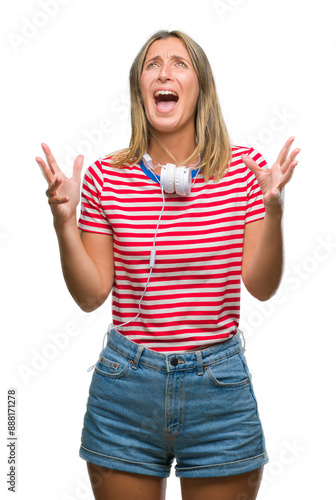 Young beautiful woman listening music wearing headphones over isolated background crazy and mad shouting and yelling with aggressive expression and arms raised. Frustration concept.