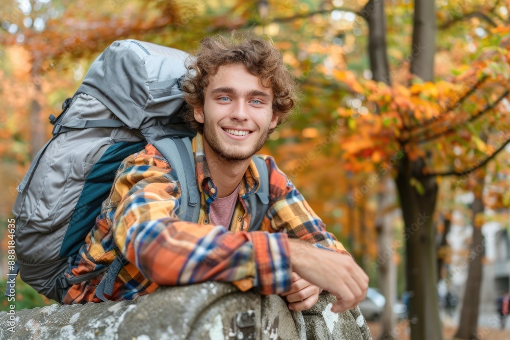 A young man with a backpack smiles brightly as he leans against a stone wall in a colorful autumn forest A young man with a backpack smiles brightly as he leans against a stone wall in a colorful autumn forest