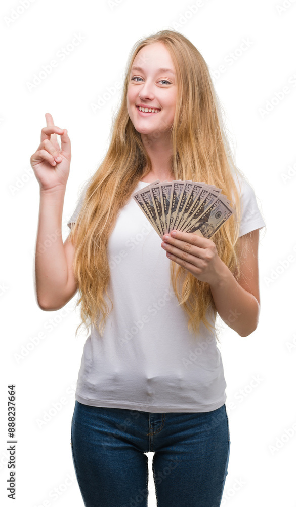 Blonde teenager woman holding dollars surprised with an idea or question pointing finger with happy face, number one Blonde teenager woman holding dollars surprised with an idea or question pointing finger with happy face, number one
