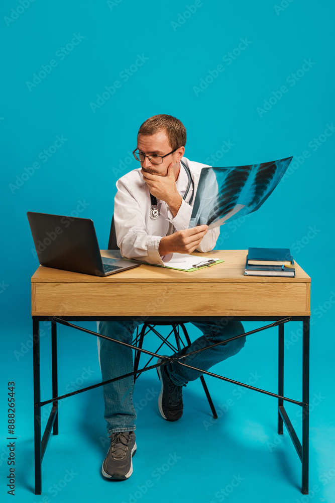 Man, doctor sitting at table, holding x ray and looking on laptop with thoughtful face against cyan background. Professional lecture. Concept of medicine, healthcare, online services, profession Man, doctor sitting at table, holding x ray and looking on laptop with thoughtful face against cyan background. Professional lecture. Concept of medicine, healthcare, online services, profession